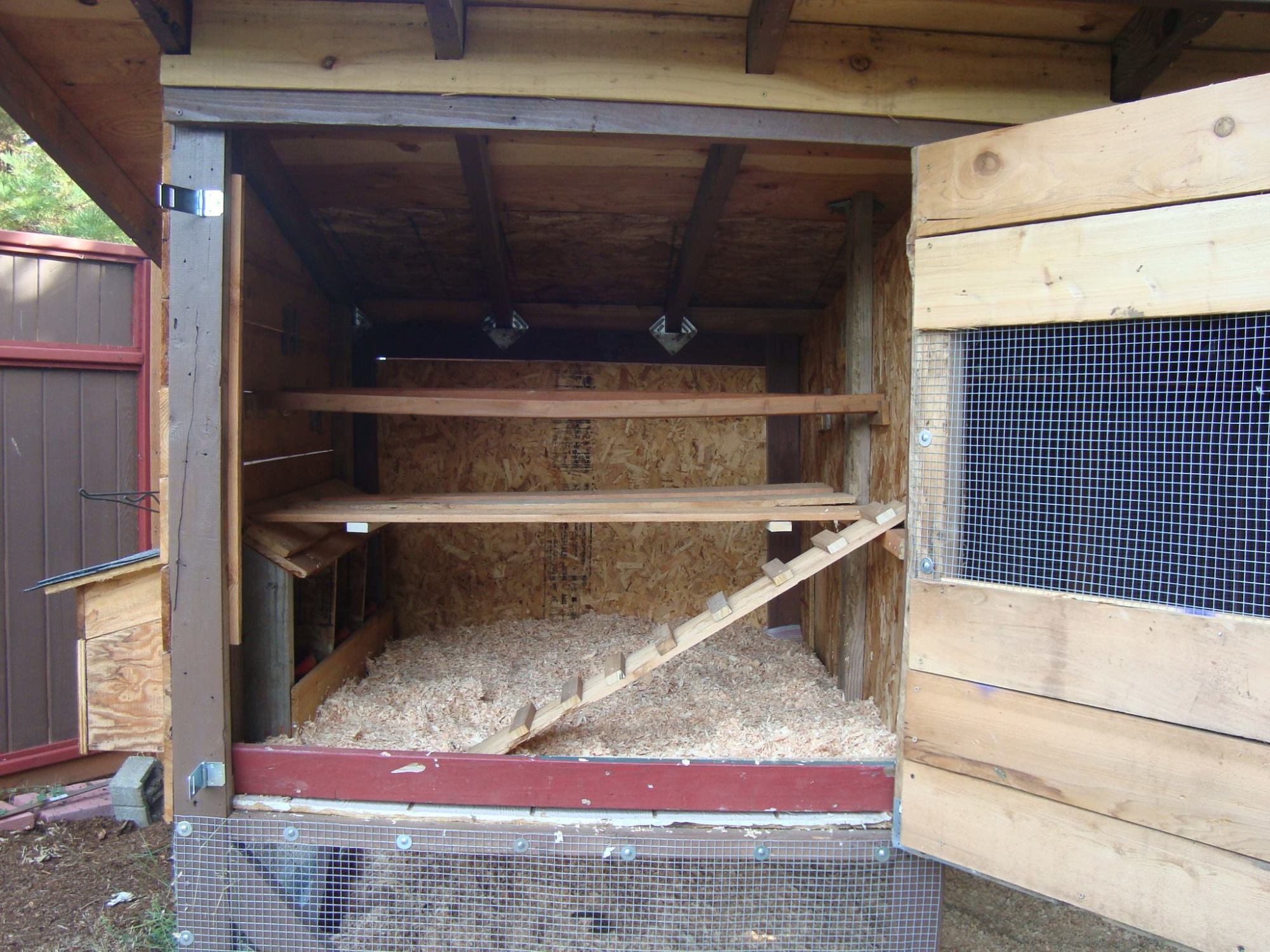 A platform beneath the roosting bars on which to place a light-weight poop board I can slide toward me for daily cleaning. Or until I find something that works better.
The floor is made of exterior siding, exterior side in, and lined with a scrap of vinyl flooring, then wood shavings. Again, end goal is to use sand in here as well.
The red board at the opening lifts out for easy raking out and while in, keeps bedding from falling out.