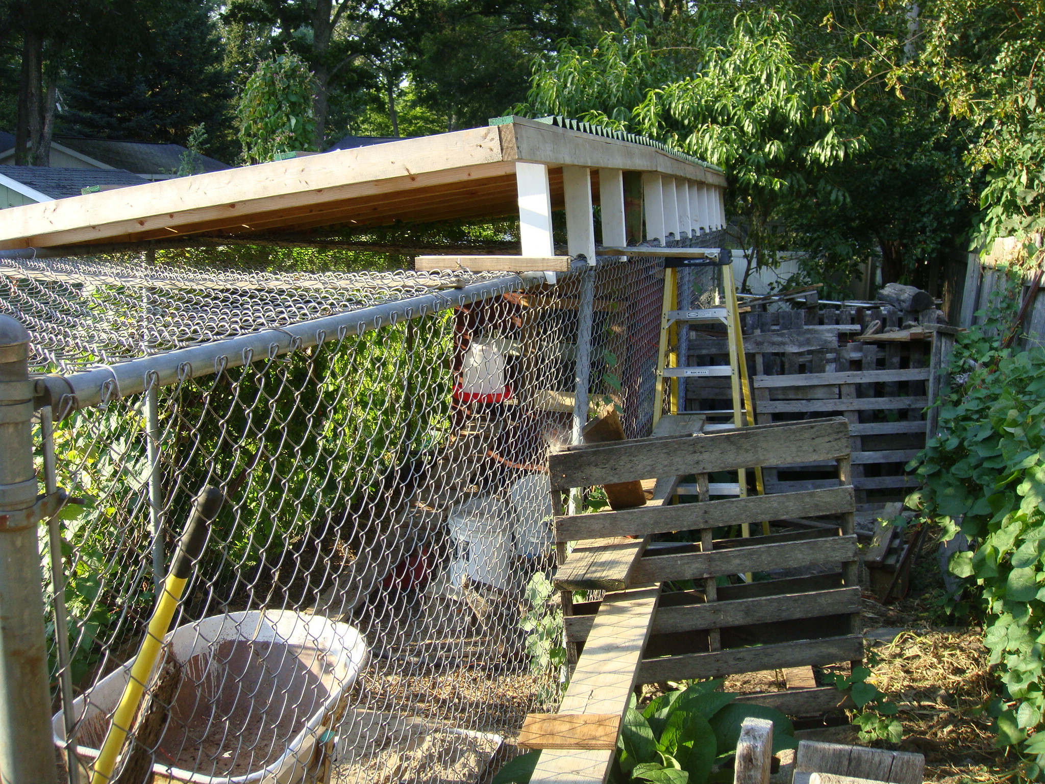 The 2 x 4 " pcs were added  (bolted) to the roofing sections we constructed in the driveway. They are the white vertical pcs about 18 " long holding the east side of the roof up. Note rounded bottom  cut out of The "risers" to fit around the pipe framework of the fencing.. Also see the pallates used as part of some scaffolding helpful in working on roof.  [ If used, be sure lumber is strong enough and fastened together solidly enough to support your wweight.... I didn't do that. ] OUCH ... Only hurt for a little while.