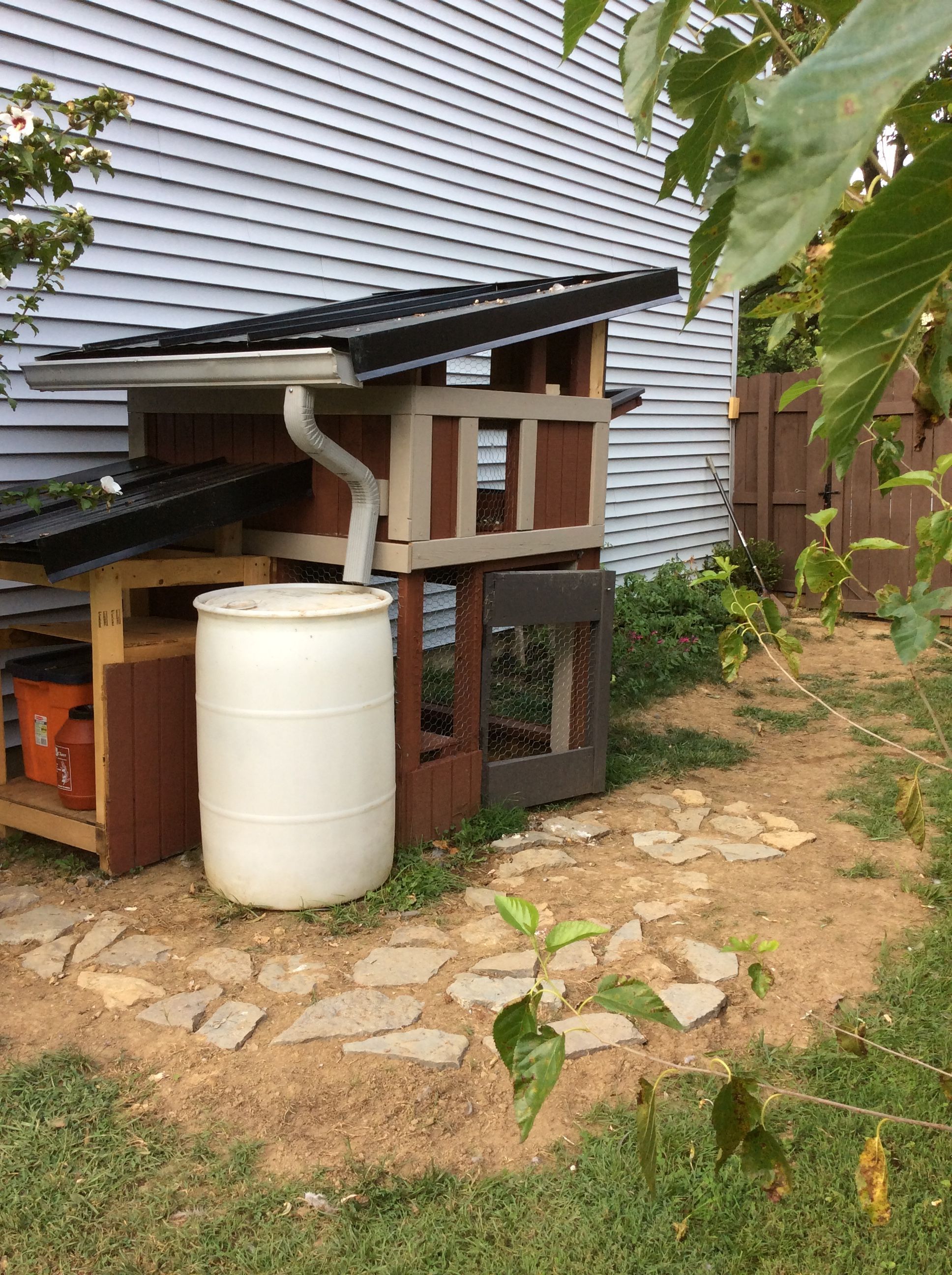 This photo was taken August 22, 2015. I added the stones around the front of the coop in hopes that snow will be easier remove from that area. 
Here, you can also see the unfinished, attached structure on the left, which is the closet. I've come to a stall on material to finish it, however ; I need screws to attach the walls and to construct the door.
 I also still need to clean the rain barrel and install a spout on the bottom of the barrel, so I can claim the water. I got the barrel from my neighbor, who was throwing it out, a few years ago. I knew I could eventually use it, so I grabbed it, and hung onto it until I had the right purpose for it. One of these weekends, soon, and before winter, I'll clean it on the inside with a pressure washer and finish it, too.