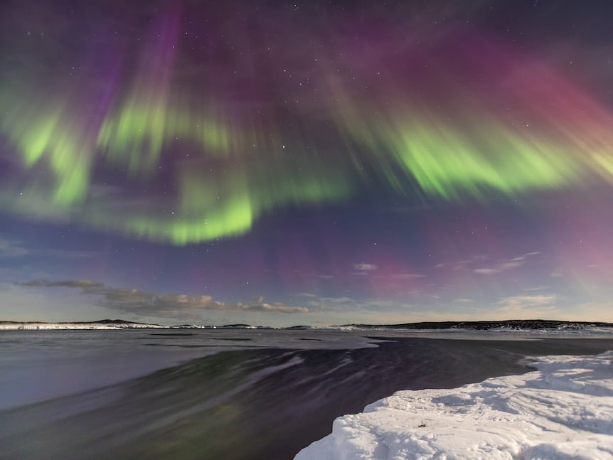 A green and pink aurora agains a dark blue sky stretches above a clear body of water in Antarctica. 